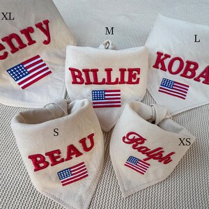 May include: Five off-white fabric pet bandanas in sizes XS, S, M, L, and XL. Each bandana features red embroidered names: Henry, Billie, Koba, Beau, and Ralph, accompanied by a small embroidered American flag patch below the name.