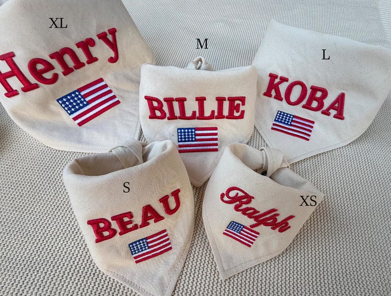 May include: Five off-white fabric pet bandanas in sizes XS, S, M, L, and XL. Each bandana features red embroidered names: Henry, Billie, Koba, Beau, and Ralph, accompanied by a small embroidered American flag patch below the name.