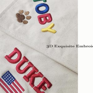 May include: Off-white fabric featuring 3D embroidery of the names TOBY and DUKE in colourful block letters. TOBY is accompanied by a brown paw print, while DUKE is paired with a small American flag. The text 3D Exquisite Embroidery is visible.