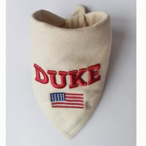 May include: A cream-coloured triangular pet bandana featuring the embroidered red text DUKE positioned above a small, rectangular embroidered American flag with red, white, and blue thread. The fabric has a visible weave and a tied top corner.