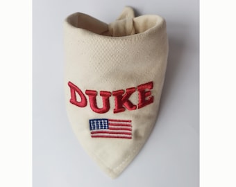 May include: A cream-colored triangular pet bandana featuring the name DUKE embroidered in bold red capital letters. Below the name, a small American flag is embroidered in red, white, and blue thread. The fabric has a visible woven texture.