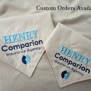 May include: Two triangular beige fabric bandanas featuring embroidered text that reads HENRY Companion Insurance Agency in light blue and dark blue thread, accompanied by a matching logo. The text Custom Orders Available is visible.