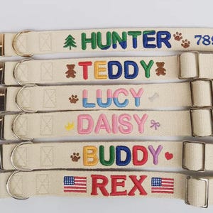 May include: Six beige fabric pet collars with silver metal buckles and D-rings. Each collar features embroidered names including HUNTER, TEDDY, LUCY, DAISY, BUDDY, and REX, accompanied by colourful icons like trees, paw prints, bears, bones, and flags.