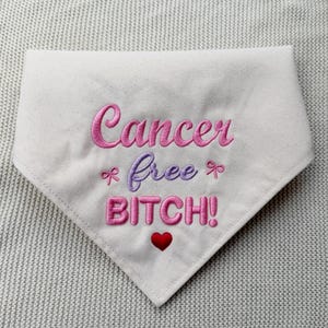 May include: A white triangular pet bandana featuring pink and purple embroidered text that reads Cancer free BITCH! with two small pink bows flanking the word free and a small red heart embroidered below the word BITCH.