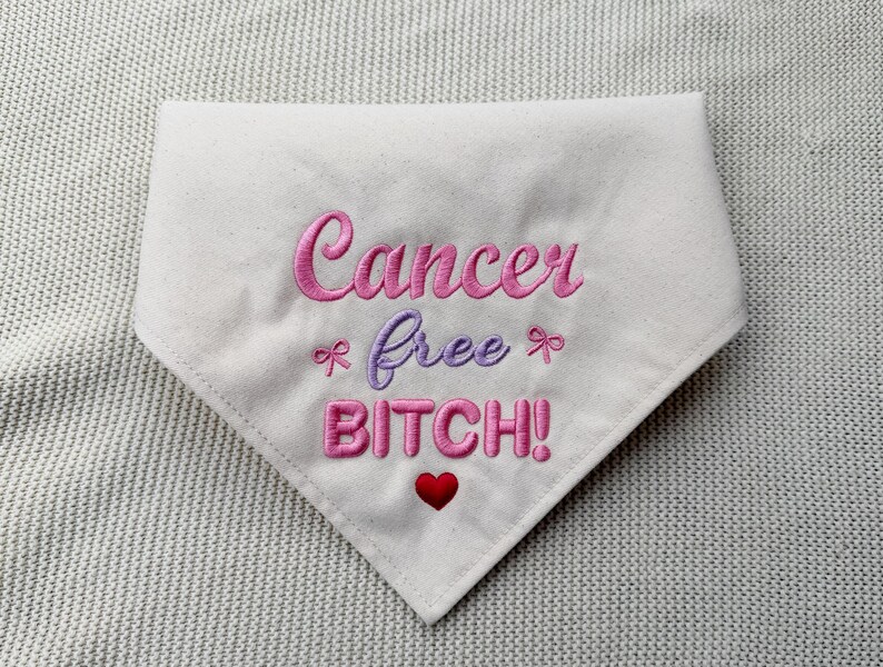 May include: A white triangular pet bandana featuring pink and purple embroidered text that reads Cancer free BITCH! with two small pink bows flanking the word free and a small red heart embroidered below the word BITCH.