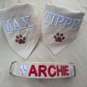 May include: Two cream-coloured triangular pet bandanas featuring embroidered white names MAX and TIPPER with brown paw prints, and one cream-coloured pet collar with the red embroidered name ARCHIE flanked by two small baseball graphics.