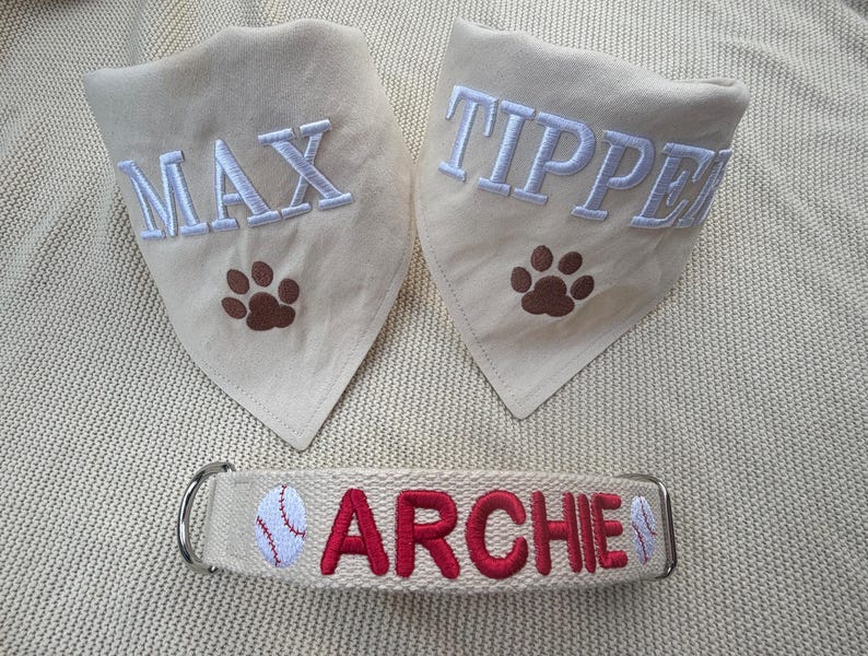 May include: Two cream-coloured triangular pet bandanas featuring embroidered white names MAX and TIPPER with brown paw prints, and one cream-coloured pet collar with the red embroidered name ARCHIE flanked by two small baseball graphics.