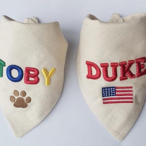 May include: Two off-white triangular pet bandanas made of canvas fabric. The left bandana features the name TOBY embroidered in green, blue, red, and yellow, with a brown paw print below. The right bandana features the name DUKE in red with an American flag.