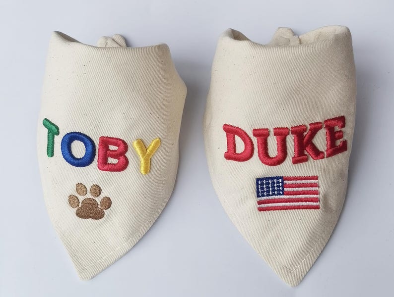 May include: Two off-white triangular pet bandanas made of canvas fabric. The left bandana features the name TOBY embroidered in green, blue, red, and yellow, with a brown paw print below. The right bandana features the name DUKE in red with an American flag.