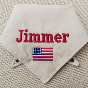 May include: An off-white triangular pet bandana featuring the embroidered name Jimmer in red capital letters. Below the name is an embroidered rectangular American flag with red, white, and blue thread. The bandana includes two metal snap fasteners.