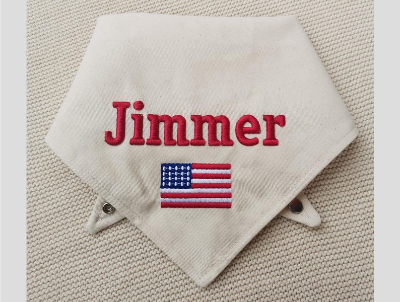 May include: An off-white triangular pet bandana featuring the embroidered name Jimmer in red capital letters. Below the name is an embroidered rectangular American flag with red, white, and blue thread. The bandana includes two metal snap fasteners.