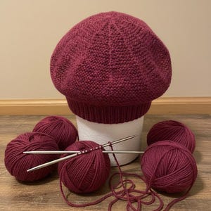 Puede incluir: Un boina de punto burdeos se encuentra sobre una forma de espuma blanca, rodeada de ovillos de lana a juego y agujas de tejer. La boina tiene un borde acanalado y una corona texturizada. El conjunto sugiere un proyecto de tejido hecho a mano.