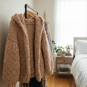 K&ouml;nnte beinhalten: Ein grob gestrickter Kapuzen-Cardigan in Hellbraun h&auml;ngt an einer Kleiderstange. Der &uuml;bergro&szlig;e Pullover hat ein dickes, strukturiertes Aussehen. Weitere Kleidungsst&uuml;cke sind auf der Stange zu sehen, was auf ein Bekleidungsthema hindeutet.