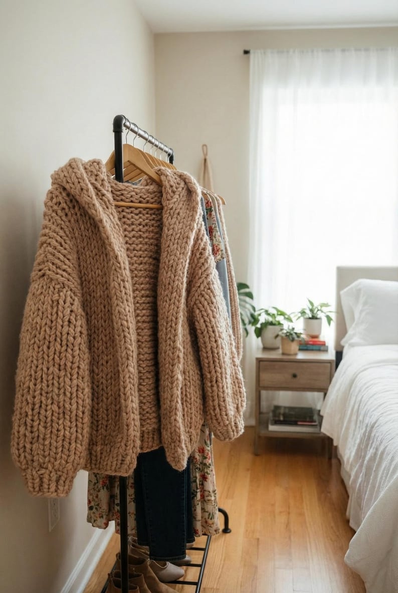 K&ouml;nnte beinhalten: Ein grob gestrickter Kapuzen-Cardigan in Hellbraun h&auml;ngt an einer Kleiderstange. Der &uuml;bergro&szlig;e Pullover hat ein dickes, strukturiertes Aussehen. Weitere Kleidungsst&uuml;cke sind auf der Stange zu sehen, was auf ein Bekleidungsthema hindeutet.
