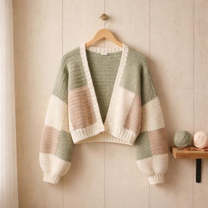 May include: A handmade, open-front crochet cardigan in a patchwork design. The sweater features a mix of colours, including sage green, cream, and blush pink. The sleeves are long and voluminous, and the cardigan is displayed on a wooden hanger.