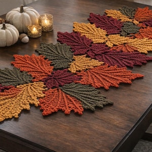May include: A crochet table runner composed of interconnected autumn leaves in shades of dark green, deep red, burnt orange, and golden yellow. The runner features a textured, leaf-shaped design that extends across the center of the wooden table.
