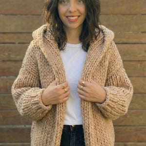 K&ouml;nnte beinhalten: Eine Frau tr&auml;gt einen beigefarbenen Kapuzen-Cardigan. Der grob gestrickte Pullover hat eine lockere Passform und eine gro&szlig;e Kapuze. Die Frau tr&auml;gt ein wei&szlig;es T-Shirt und dunkle Jeans. Der Hintergrund ist ein Holzzaun.