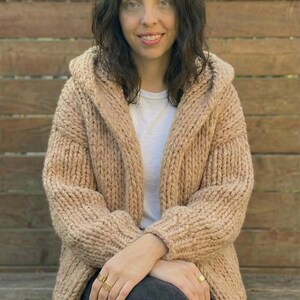 K&ouml;nnte beinhalten: Eine Frau tr&auml;gt eine beige Kapuzenstrickjacke. Der grob gestrickte Pullover hat eine lockere Passform und ist vorne offen. Sie tr&auml;gt ein wei&szlig;es T-Shirt und dunkle Jeans. Der Hintergrund ist ein Holzzaun.