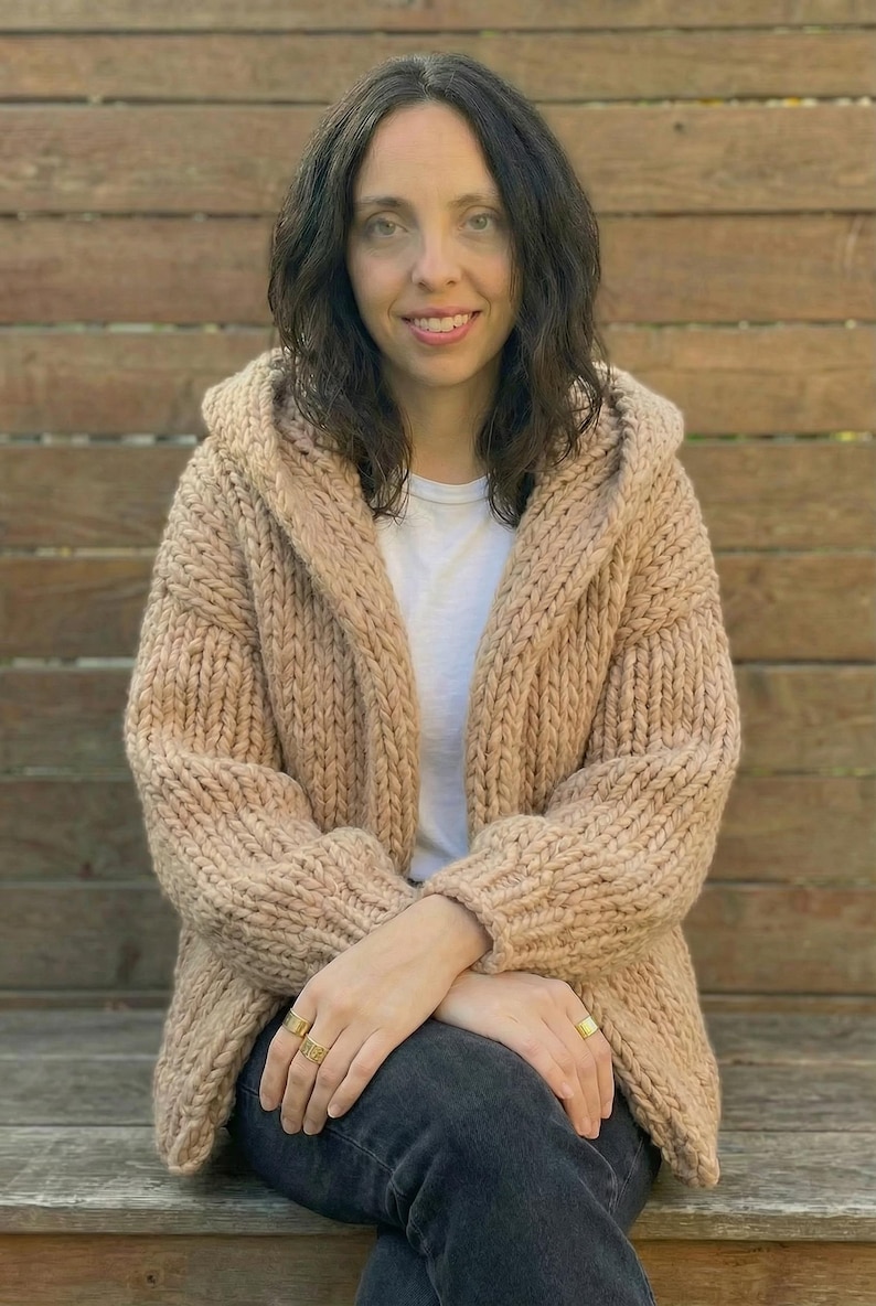 K&ouml;nnte beinhalten: Eine Frau tr&auml;gt eine beige Kapuzenstrickjacke. Der grob gestrickte Pullover hat eine lockere Passform und ist vorne offen. Sie tr&auml;gt ein wei&szlig;es T-Shirt und dunkle Jeans. Der Hintergrund ist ein Holzzaun.