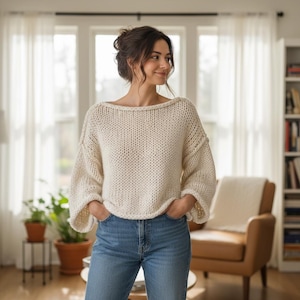 May include: A cream-colored, chunky knit sweater with a wide boat neckline and loose, long sleeves. The sweater features a thick, textured yarn construction with visible stitching and a relaxed fit, paired with classic blue denim jeans.