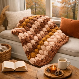 May include: A handmade, textured blanket in shades of brown, tan, and peach, draped over a window seat. The blanket features a raised, bobble-stitch pattern. A cozy, warm home decor item.