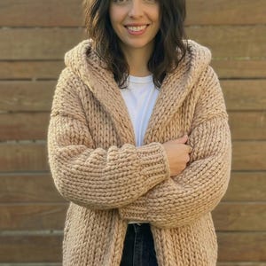 K&ouml;nnte beinhalten: Beige Kapuzen-Cardigan mit grobem Strickdesign. Der offene Pullover hat lange &Auml;rmel und eine lockere Passform. Das Model tr&auml;gt ein wei&szlig;es T-Shirt und dunkle Jeans. Der Hintergrund ist ein Holzzaun.