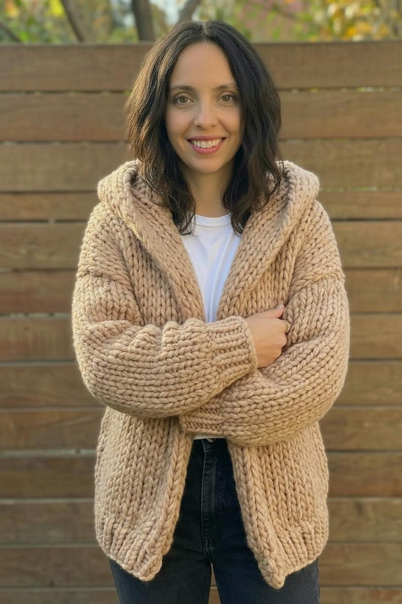 K&ouml;nnte beinhalten: Beige Kapuzen-Cardigan mit grobem Strickdesign. Der offene Pullover hat lange &Auml;rmel und eine lockere Passform. Das Model tr&auml;gt ein wei&szlig;es T-Shirt und dunkle Jeans. Der Hintergrund ist ein Holzzaun.