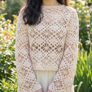 May include: Cream-colored crochet top with long sleeves. The top features a floral pattern throughout the design. The crochet work is open, creating a delicate and airy look. The top is worn over a lighter-colored garment.