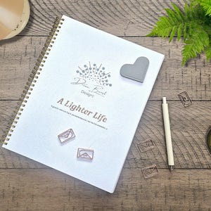 May include: A white spiral-bound notebook with the text "Dream Burst Design" and "A Lighter Life" on the cover. A grey heart-shaped magnet, a white pen, and three rose gold paper clips are arranged on a wooden surface.