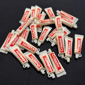 May include: A collection of miniature Colgate toothpaste tubes. Each tube is white with a red rectangular label bearing the Colgate logo. The tubes are scattered on a black surface. The text "NET WT. 1.3 OZ" is visible.