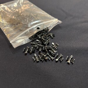 Puede incluir: Una colección de pequeños dijes metálicos oscuros con forma de hormiga que salen de una bolsa de plástico transparente resellable. Cada dije presenta un cuerpo segmentado y seis patas delgadas, con un acabado liso y reflectante.