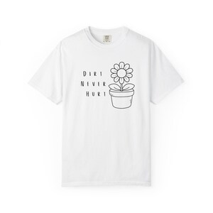 Puede incluir: Camiseta blanca con un dibujo negro de una flor en maceta y el texto "DIRT NEVER HURT". El diseño es sencillo y limpio, ideal para uso diario. La camiseta está hecha de un material suave y cómodo.