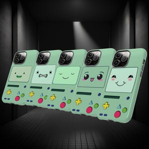 Puede incluir: Cinco fundas verdes para teléfono con caras de dibujos animados. Cada funda presenta una expresión diferente en una pantalla blanca, con botones coloridos y un mando en forma de cruz. Las fundas están diseñadas para parecerse a una consola de juegos portátil retro.
