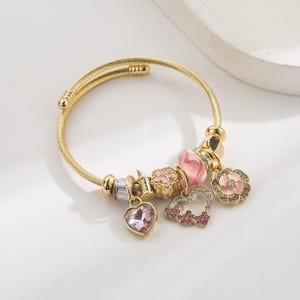 May include: Gold-coloured bracelet with various charms. Charms include a pink heart, a pink and gold flower, a pink heart-shaped charm with pink gems, and a pink bead. The bracelet is on a white surface.