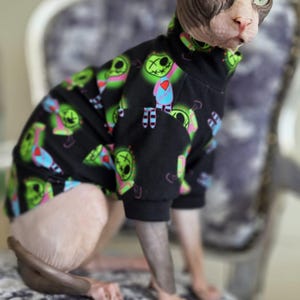 Puede incluir: Un suéter negro para mascota con cuello alto y mangas largas, que presenta un estampado gráfico repetitivo de personajes de dibujos animados en verde, azul y rosa con detalles cosidos. La prenda la lleva un gato sin pelo de orejas grandes.