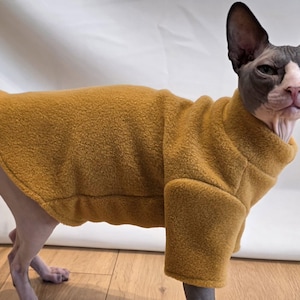 May include: A Sphynx cat wearing a mustard-coloured fleece jacket. The cat has a grey and pink face with large ears and is standing on a wooden floor. The jacket covers the cat's body and has sleeves.