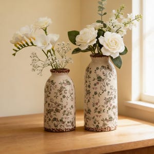Puede incluir: Dos jarrones de cerámica con motivos florales y bordes marrones sostienen flores blancas. Los jarrones son cilíndricos y tienen una base color crema con diseños florales verdes y marrones. Están sobre una superficie de madera.