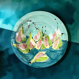 May include: A round ceramic plate featuring a hand-painted watercolor illustration of several pears. The pears are rendered in shades of light green and soft pink with dark black outlines, set against a teal and light blue background.