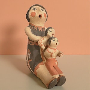Puede incluir: Una figura de arcilla pintada a mano que representa a un adulto sentado con dos figuras más pequeñas adheridas al frente. Las figuras presentan detalles pintados en color beige, negro y terracota, incluyendo rasgos faciales y patrones.