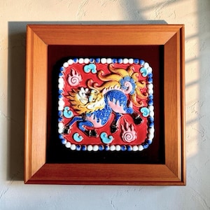 May include: A square ceramic tile featuring a relief of a mythical creature with a blue body and yellow mane, set against a red background. The tile is bordered by small white and blue spheres and is encased in a simple wooden frame.