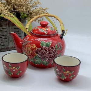 Pode incluir: Uma chaleira de cerâmica vermelha e duas chávenas a condizer. A chaleira apresenta uma pega de bambu e um design floral em tons de amarelo, verde e roxo. As chávenas têm um padrão floral semelhante. O conjunto é para chá.