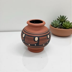 Puede incluir: Un pequeño jarrón de terracota de forma redondeada con acabado mate, que presenta bandas negras horizontales y un patrón repetitivo de formas ovaladas de color crema y pequeños puntos alrededor de la sección media, con borde ancho.