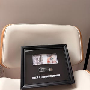 Op de afbeelding: Een ingelijst kunstwerk met een zwart frame, met een stapel van $100 biljetten en een autosleutelhanger op een zwarte achtergrond. De tekst "IN CASE OF EMERGENCY BREAK GLASS" staat eronder.