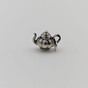 Op de afbeelding: Een kleine, zilverkleurige theepotbedel met een afgeronde body en een gebogen tuit. De bedel heeft een deksel en een handvat en is bevestigd aan een kleine ring. De theepot is waarschijnlijk van metaal en staat tegen een witte achtergrond.