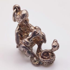 Könnte beinhalten: Ein silberfarbener Metallanhänger, der eine Figur mit Turban darstellt, die eine Flöte spielt, während eine Schlange aus einem runden Korb aufsteigt. Der Anhänger hat eine texturierte, verwitterte Oberfläche und einen kleinen Ring zur Befestigung.
