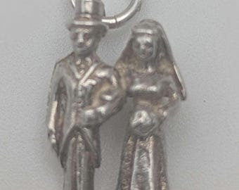 May include: A silver-toned metal charm depicting a couple in wedding attire. The figure on the left wears a top hat and suit, while the figure on the right wears a long gown and veil. A circular jump ring is attached to the top of the charm.