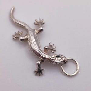 Peut inclure: Un pendentif en métal argenté en forme de lézard avec un corps texturé aux écailles visibles, quatre pattes écartées, une longue queue courbée et un petit anneau circulaire fixé à la tête pour le suspendre, sur un fond clair uni.