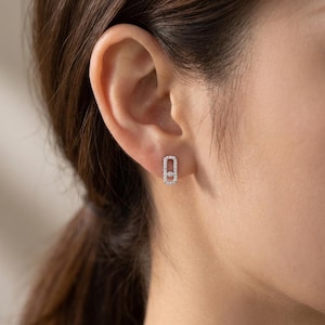 May include: A close-up of a person's ear with a small, rectangular earring. The earring is silver-toned and encrusted with small, clear gemstones. A larger, single gemstone is set in the center of the earring.