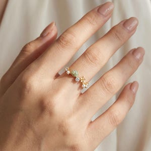 Puede incluir: Un anillo dorado con un diseño delicado de pequeñas perlas, cristales transparentes y una piedra preciosa verde claro. El anillo se lleva en un dedo, mostrando sus intrincados detalles y su estilo elegante. El diseño del anillo recuerda elementos florales o naturales.