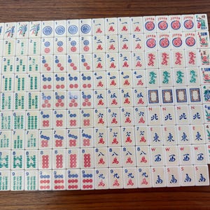 Puede incluir: Un juego completo de fichas de Mahjong de color marfil, dispuestas sobre una superficie de madera. Las fichas presentan varios símbolos, números y caracteres chinos en rojo, verde y azul. Algunas fichas tienen la palabra "Joker" y "Win".